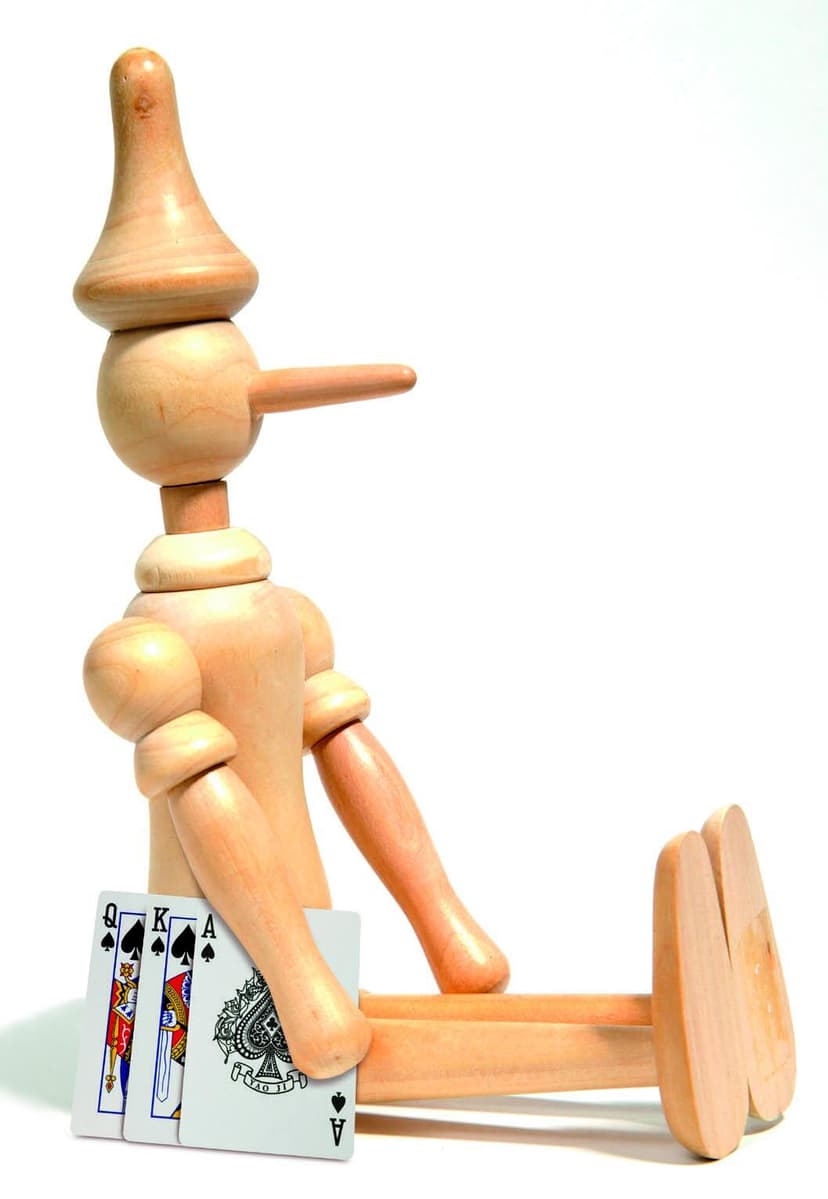 Hölzerne Pinocchio-Marionette mit langer Nase, davor Dame, König und Ass Pik als Spielkarten